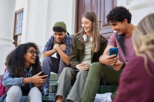 Five teens smile as they interact and share content on their phones on the school steps